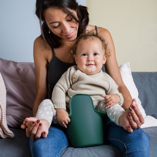 Eine Frau sitzt auf einer Couch, lächelt und hält ein Baby auf dem Schoß, das einen beigen Pullover trägt und in einem grünen Kindersitz sitzt, während es das Easy Pisi Easypisi - Das Windelfreitöpfchen in der Hand hält. Im Hintergrund sind Kissen angeordnet.