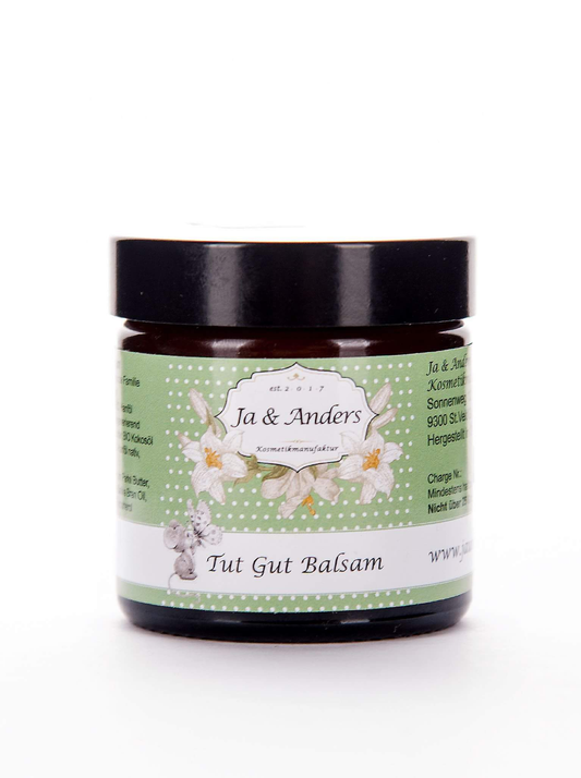 Ein dunkler Glastiegel mit schwarzem Deckel, beschriftet mit Ja & Anders Tut Gut Balsam 60g von Ja&Anders, zeigt ein grünes Etikett mit weißen Blumen und einem deutschen Text, der vegane Pflege und wasserfreien Pflegebalsam mit Bio Hanföl hervorhebt.