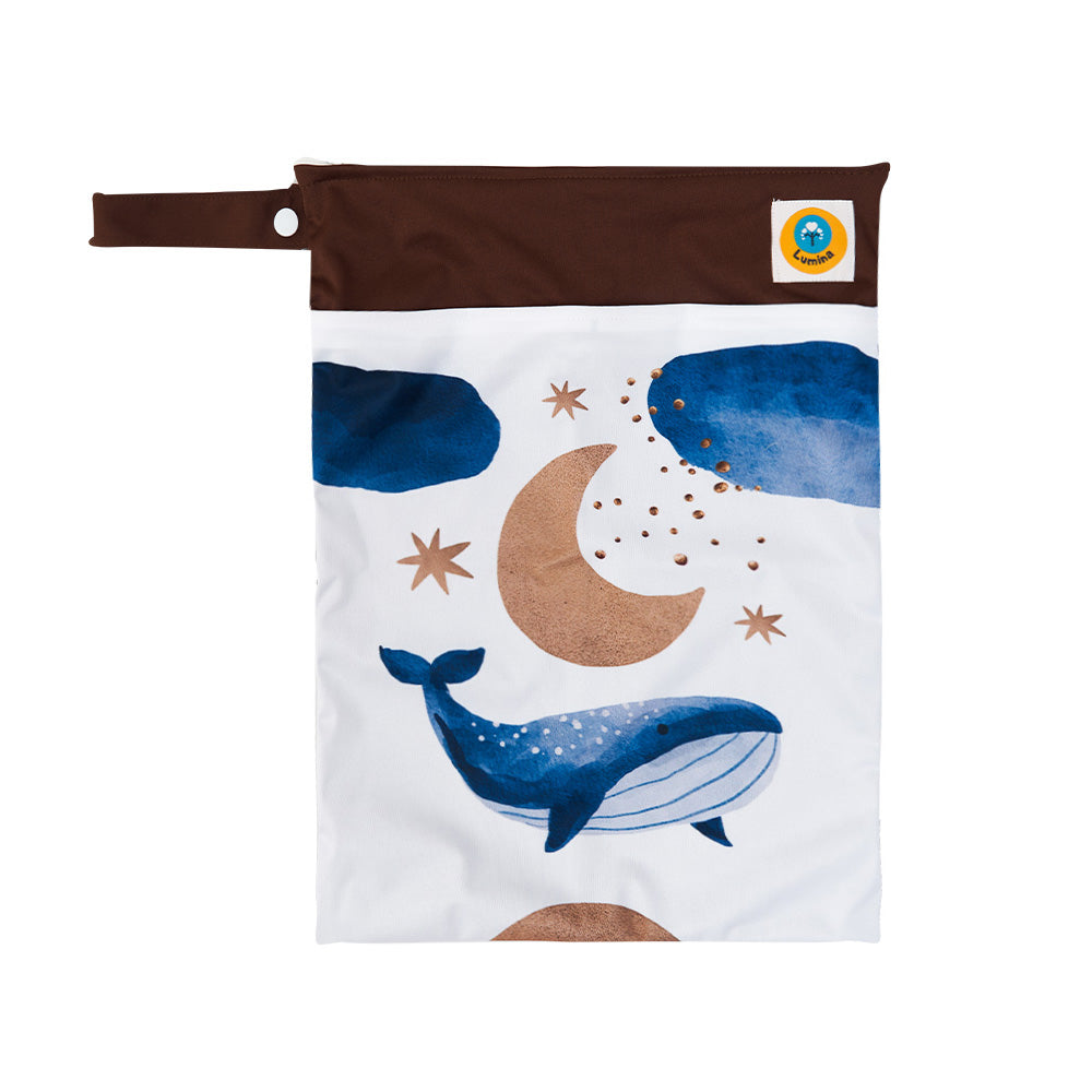 Die Lumina Doppel Wetbag/Nasstasche mit 2 Fächern big Print ist wiederverwendbar, mit braunem Deckel, Schnappgriff, blauem Wal, marineblauen Wolken, goldener Mondsichel und Sternen auf weißem Grund sowie einem runden Logoaufnäher oben rechts. Ideal für Stoffwindeln oder schmutzige Kleidung.