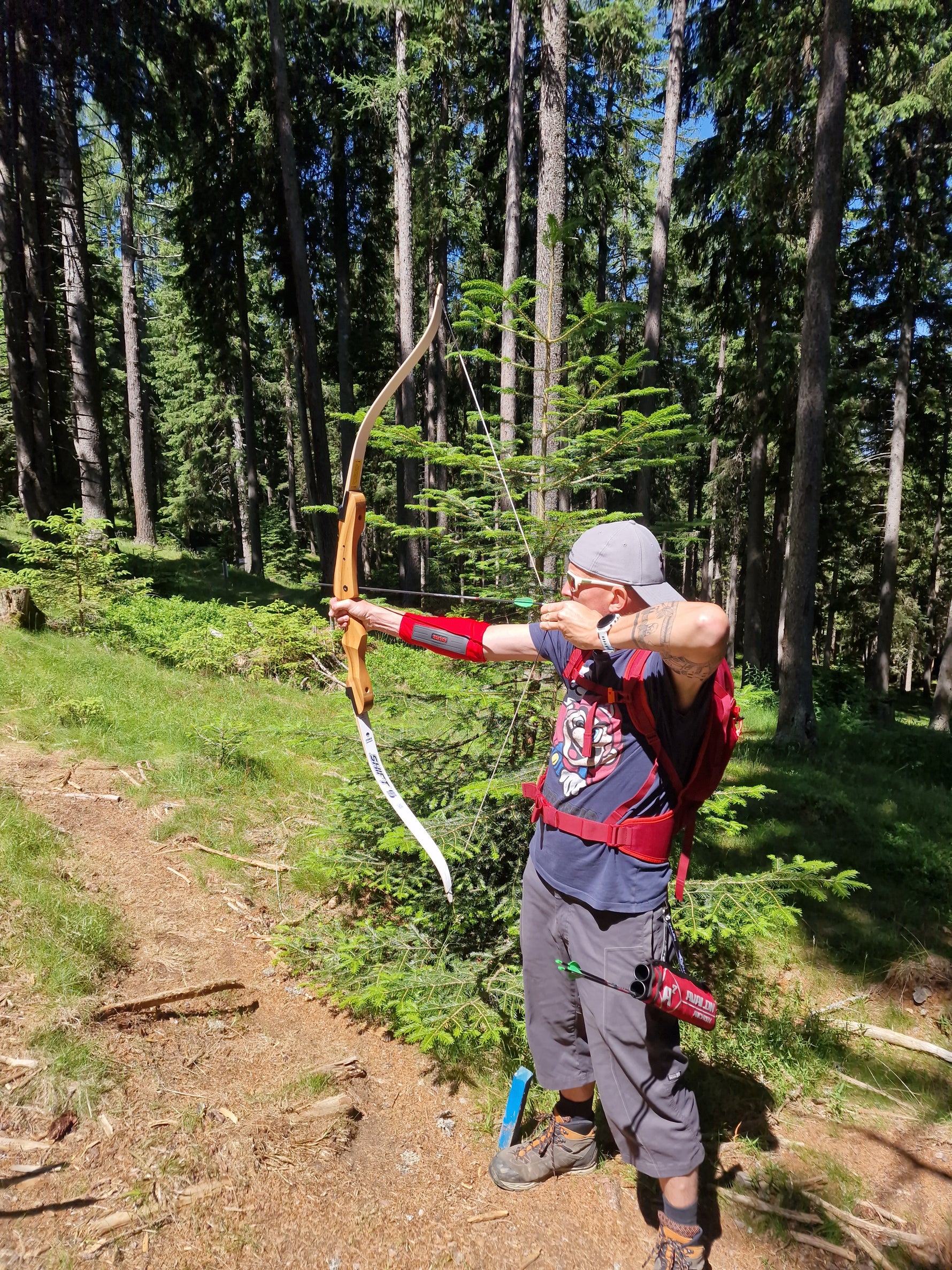 Eine Person in Outdoor-Kleidung und mit Mütze spannt einen Holzbogen und zielt auf einen Pfeil in einem sonnenbeschienenen Wald, der von hohen Bäumen und grünem Laub umgeben ist.