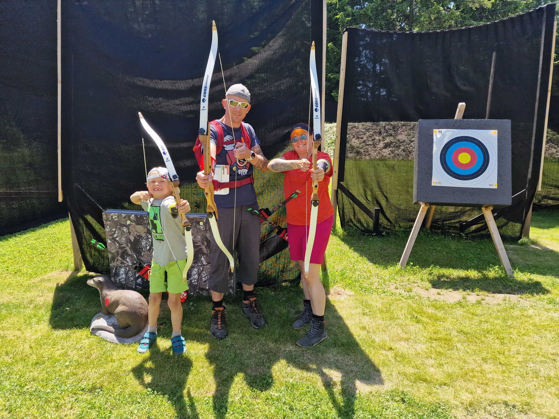 Drei Personen, zwei Erwachsene und ein Kind, posieren im Freien mit Pfeil und Bogen und zielen auf die Kamera. Sie stehen im Gras vor einer Zielscheibe und einem schwarzen Hintergrund unter einem sonnigen Himmel. Alle lächeln und sind sportlich gekleidet.