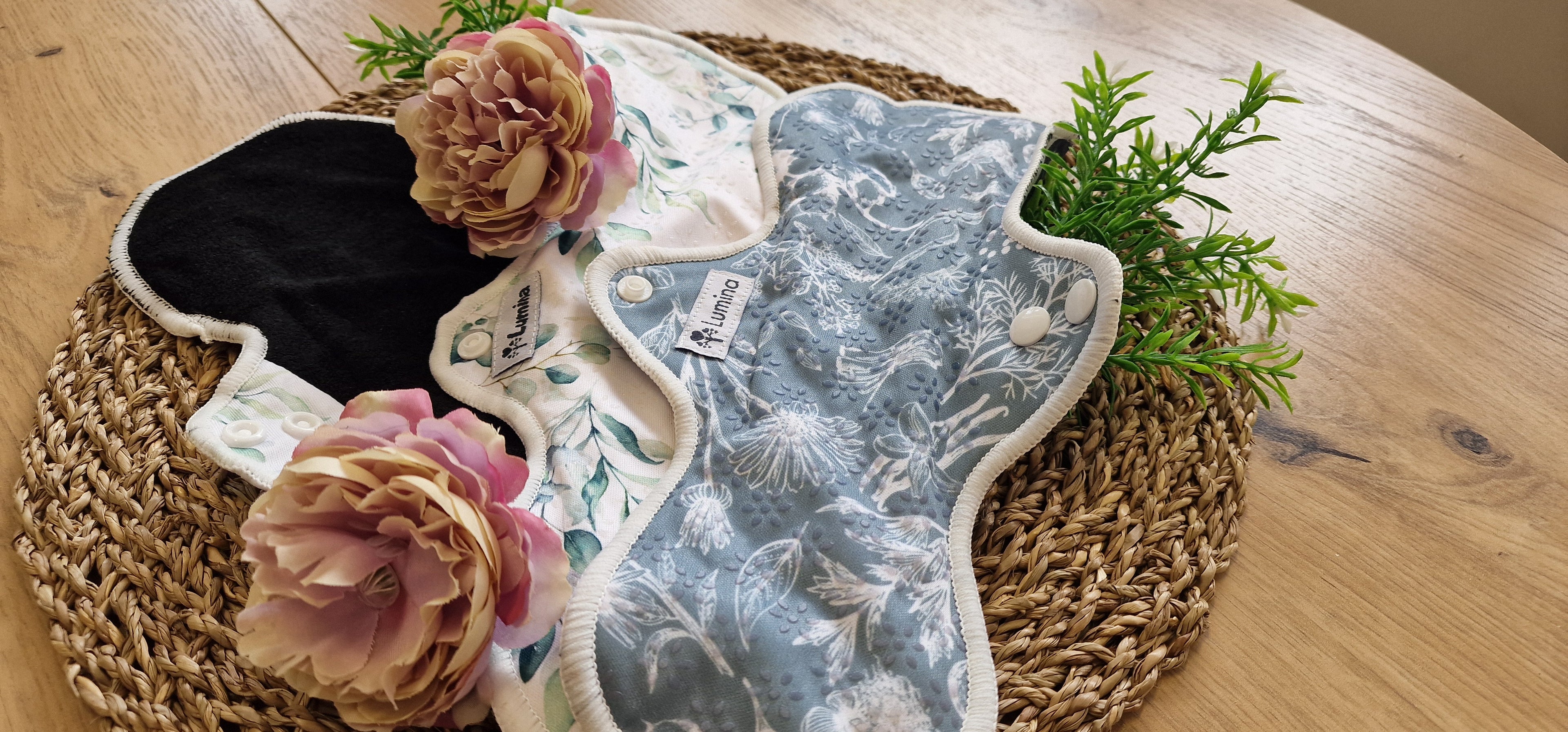 Zwei wiederverwendbare Menstruationsbinden aus Stoff mit Blumenmuster liegen auf einem gewebten Tischset, das mit zwei rosa Kunstblumen und grünem Laub verziert ist, auf einem Holztisch.