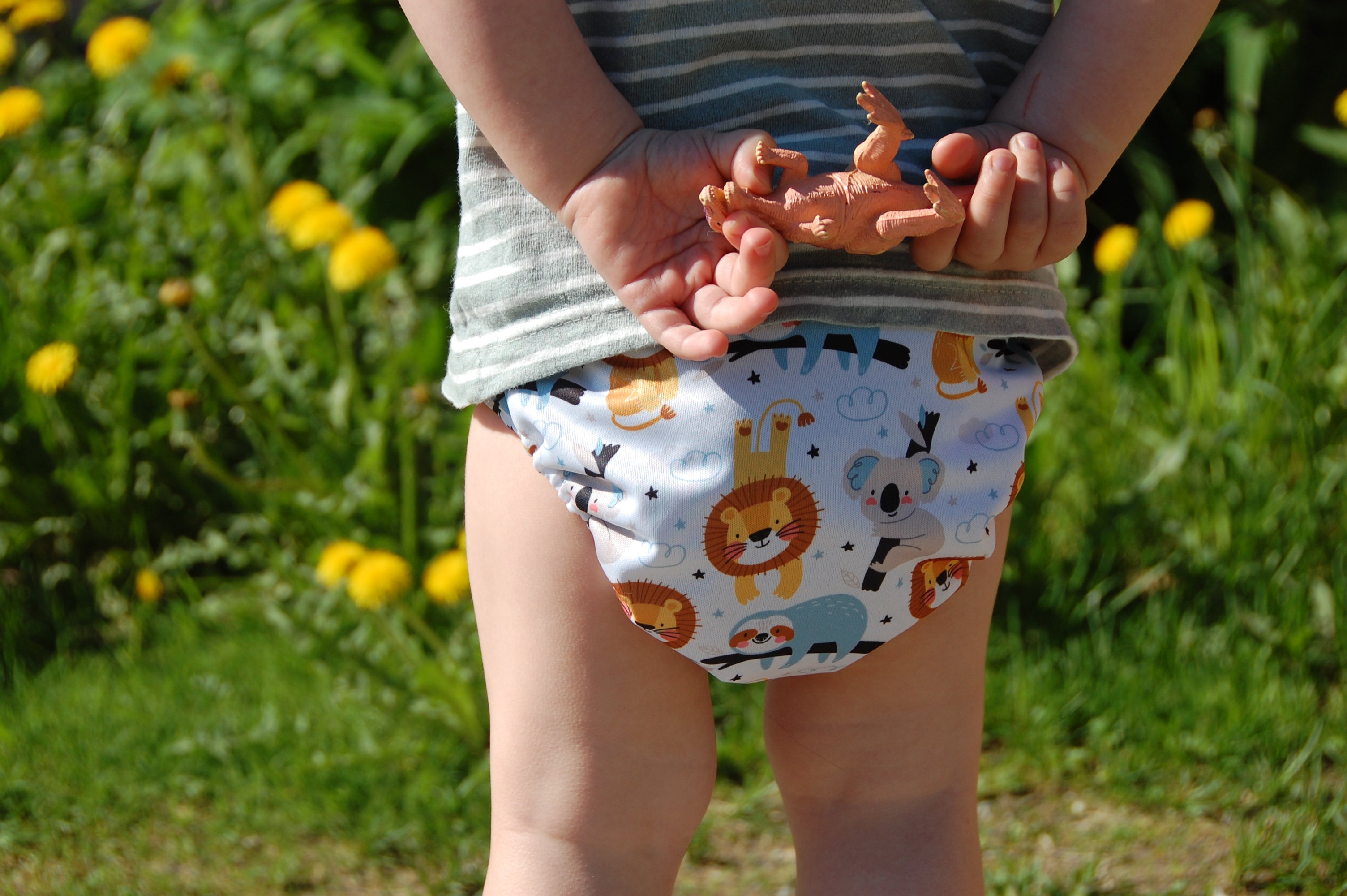 Ein Kleinkind in einem gestreiften Hemd und einer Windel mit Tiermotiven steht im Freien und hält einen kleinen Spielzeugdinosaurier hinter seinem Rücken. Im Hintergrund sind gelbe Blumen und Grünpflanzen zu sehen.