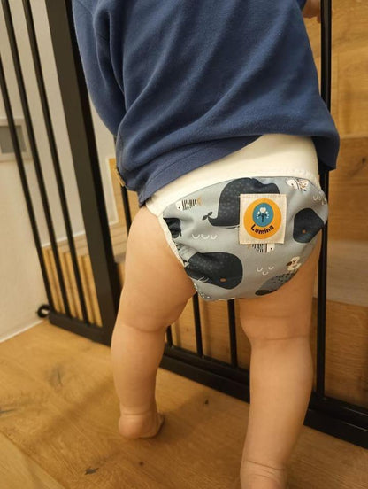 Ein Baby in einem blauen Hemd und einer Lumina Pull-me-Up All in One Windel One-Size aus GOTS zertifizierter Bio-Baumwolle mit Wal-Muster steht an einer Holztreppe und hält sich am schwarzen Sicherheitsgitter fest.