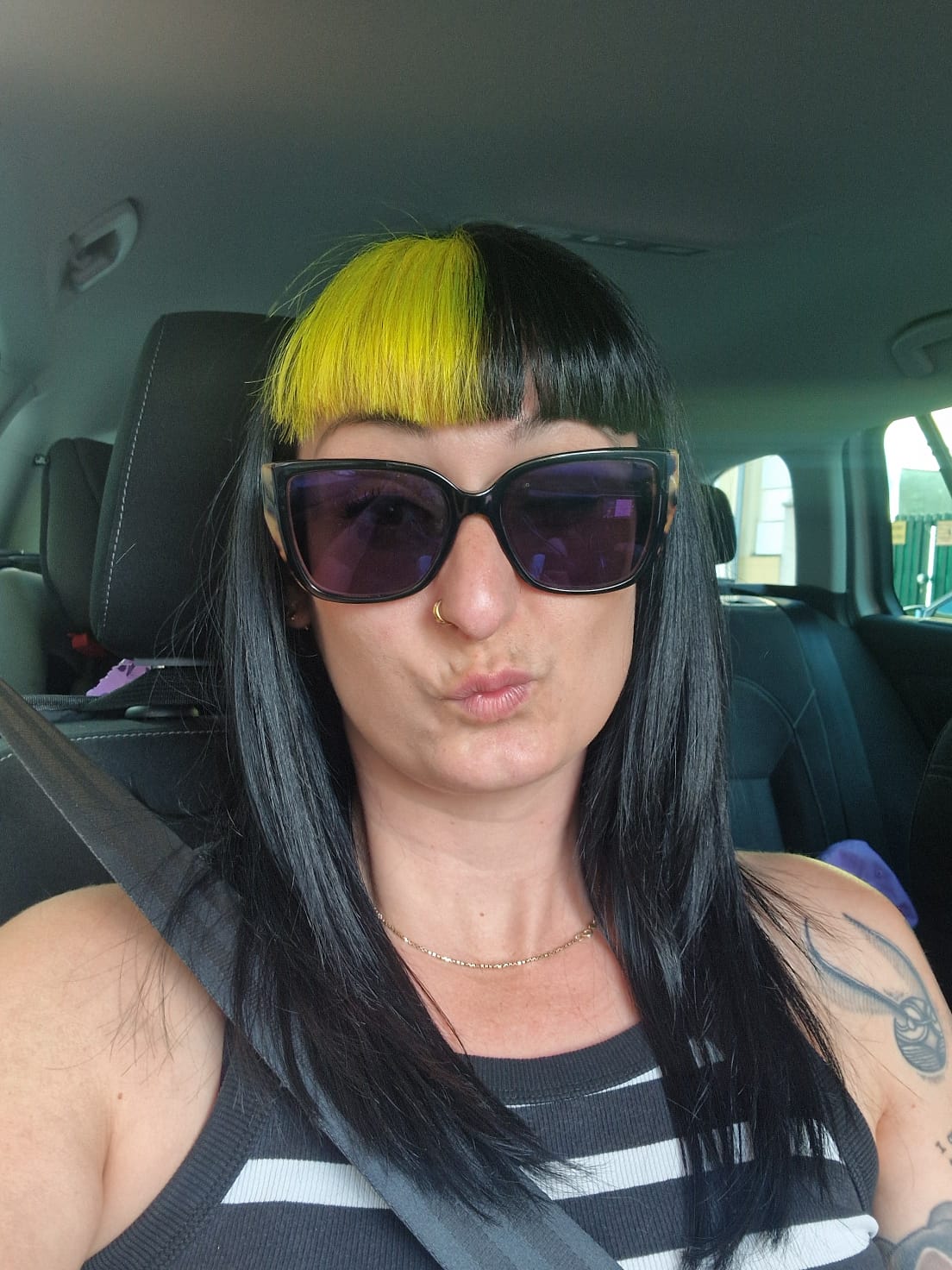 Eine Frau mit langen schwarzen Haaren und einer gelben Strähne im Pony sitzt in einem Auto, trägt eine große Sonnenbrille, ein gestreiftes Tanktop und einen Sicherheitsgurt und macht ein Entengesicht.