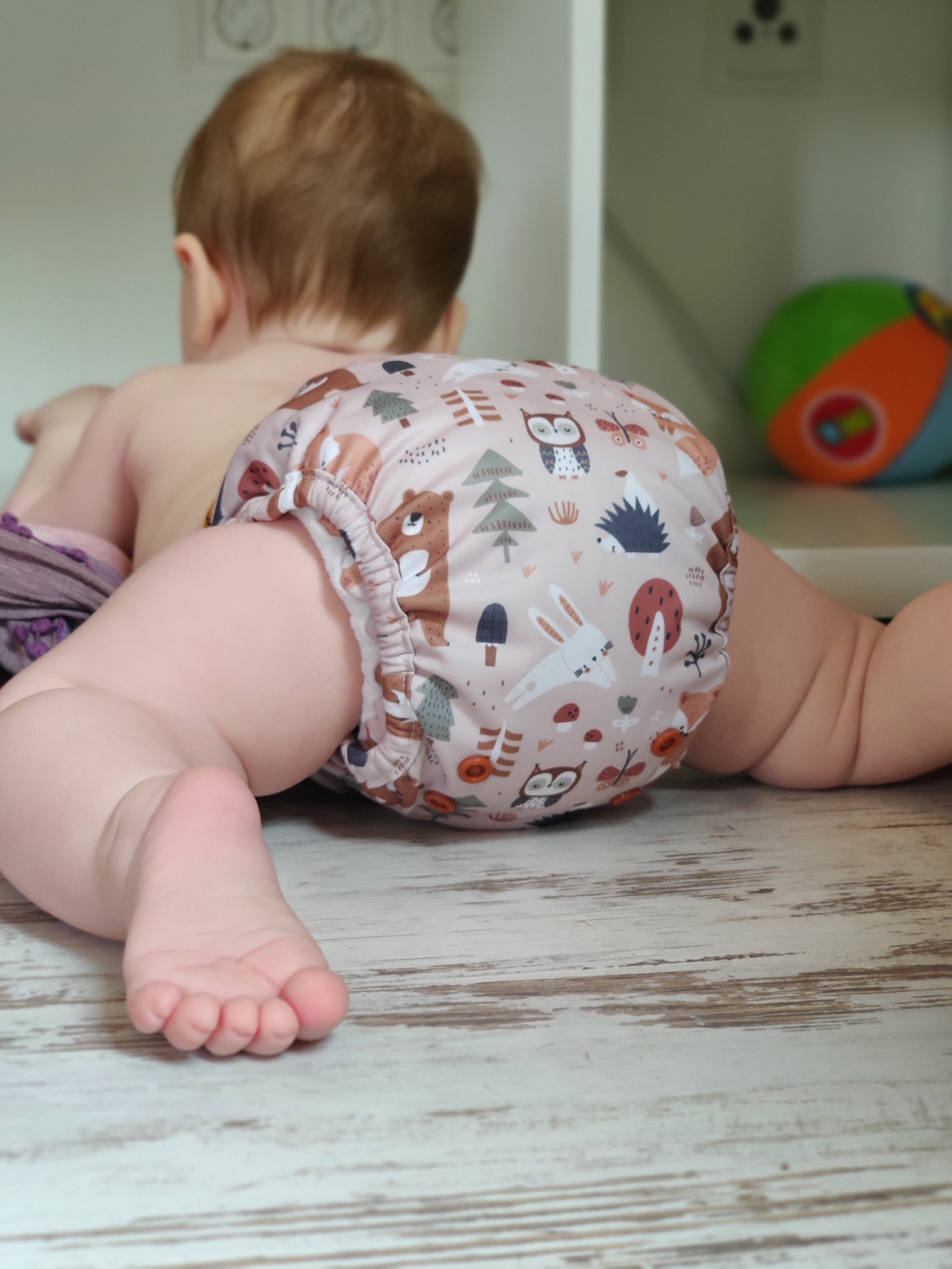 Ein Baby, das eine bunte Windel mit Tiermotiven trägt, liegt auf dem Bauch auf einem Holzboden und schaut von der Kamera weg. Im Hintergrund ist ein bunter Ball zu sehen.