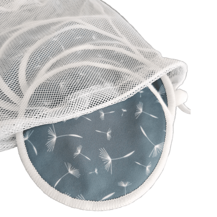 Die Lumina Stilleinlagen 3 Paar, wiederverwendbare blaue Pads mit weißem Pusteblumenaufdruck, sind teilweise in einem weißen Netzwäschesack abgebildet, beide auf einer weißen Fläche.