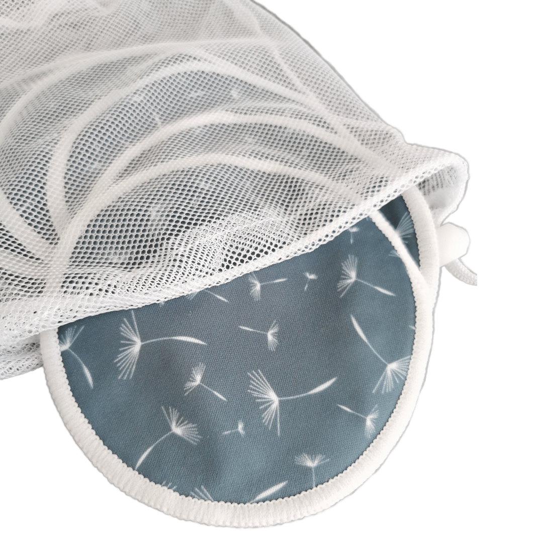 Ein weißer Wäschesack aus Mesh, teilweise geöffnet, enthüllt einen runden blauen Lumina Stilleinlagen 3 Paar Block mit einem Pusteblumenmuster und Saugkern aus Hanf im Inneren, vor einem weißen Hintergrund.