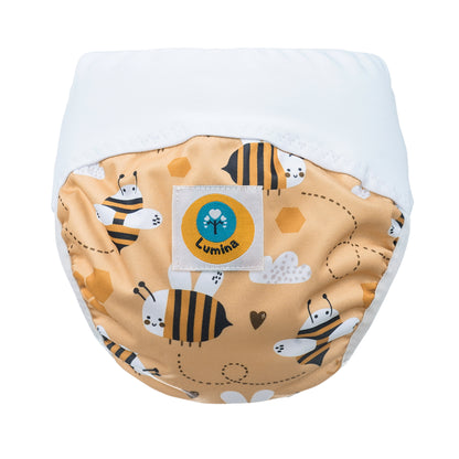 Die Lumina Pull-me-Up All in One Windel One-Size hat einen weißen Bund, ein gelbes Paneel mit Bienen, Waben, Herzen und Blumen bedruckt, ist aus GOTS-zertifizierter BioBaumwolle und hat einen blau/gelben runden Lumina-Logoaufnäher auf der Vorderseite.