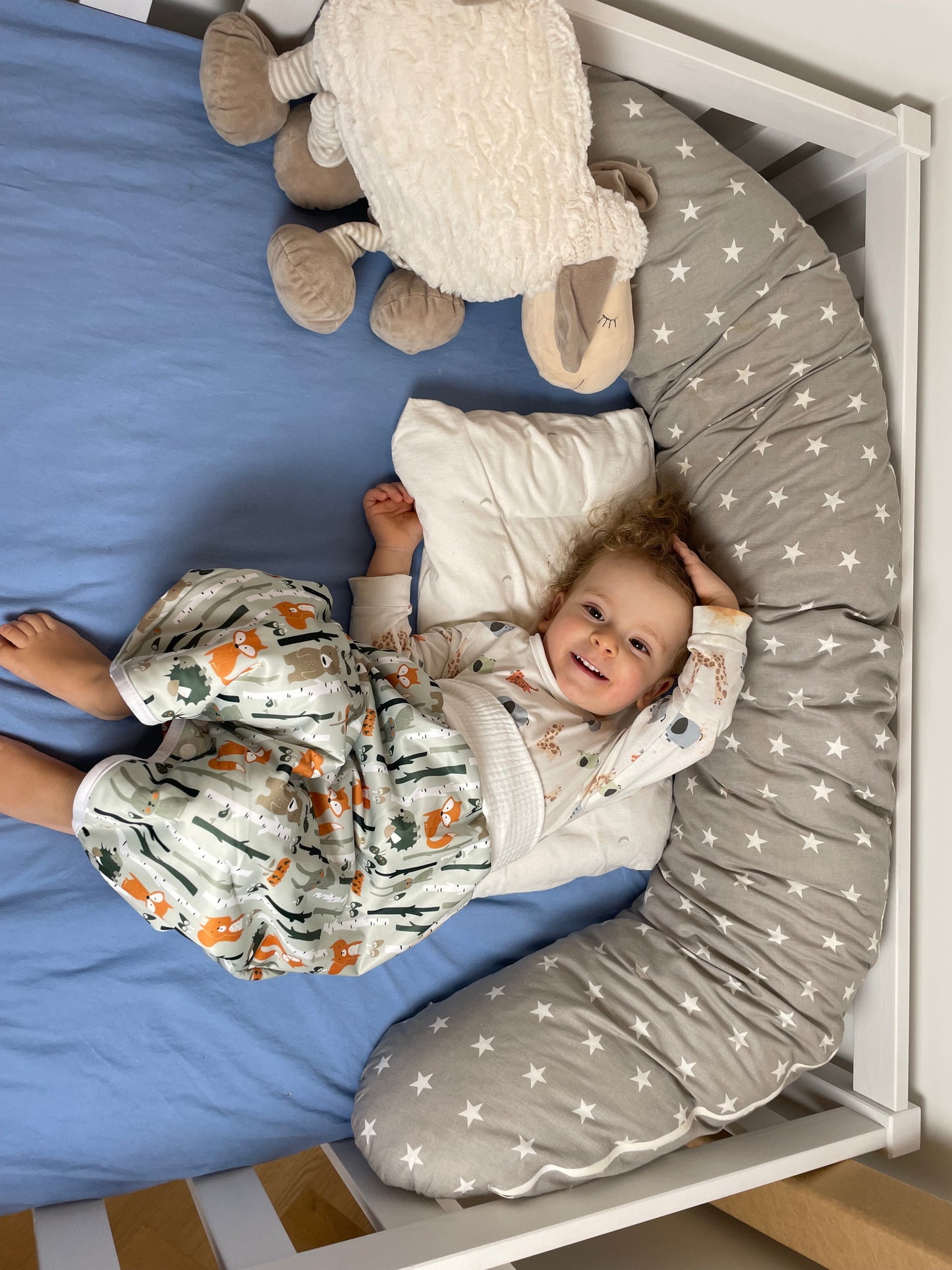 Ein lächelndes Kleinkind liegt auf einem blauen Laken in einem Kinderbett und trägt einen Schlafanzug mit Tieren darauf. Um sie herum liegen ein weißes Kissen, ein Plüschschaf und ein graues, mit Sternen gemustertes Kissen.