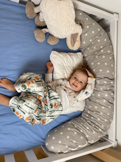 Ein lächelndes Kleinkind liegt auf einem blauen Laken in einem Kinderbett und trägt einen Schlafanzug mit Tieren darauf. Um sie herum liegen ein weißes Kissen, ein Plüschschaf und ein graues, mit Sternen gemustertes Kissen.