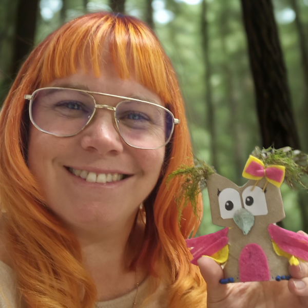Eine lächelnde Person mit orangefarbenen Haaren und Brille hält eine handgefertigte Eule mit rosafarbenen Flügeln und einer gelben Schleife in der Hand und steht in einem Wald mit hohen Bäumen im Hintergrund.