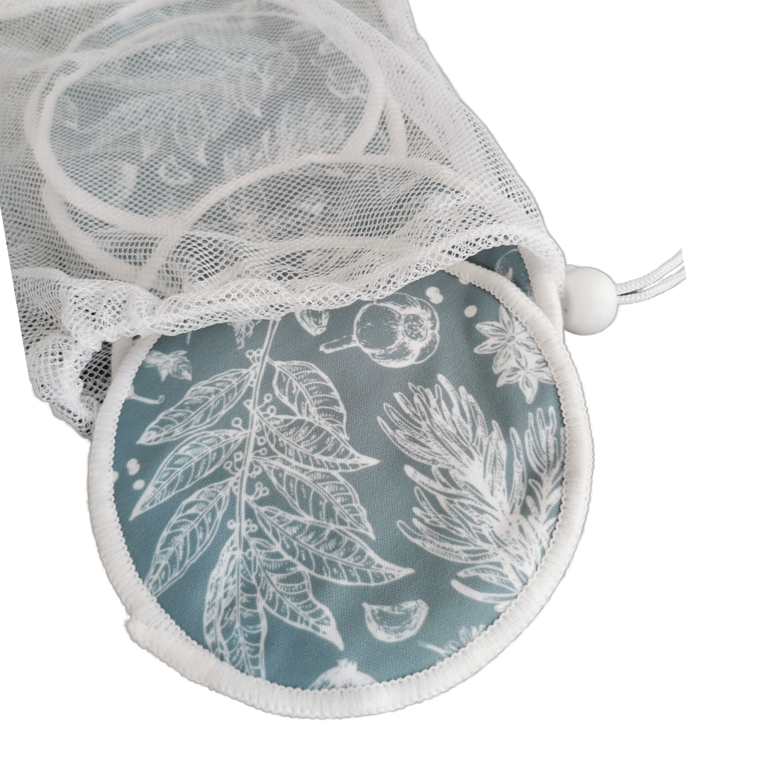 Ein Netzbeutel mit Kordelzug, der teilweise geöffnet ist, enthüllt Lumina Stilleinlagen 3 Paar - runde, wiederverwendbare Baumwollpads mit weißen botanischen Blatt- und Fruchtmustern auf blauem Grund, die jeweils einen saugfähigen Hanfkern haben.